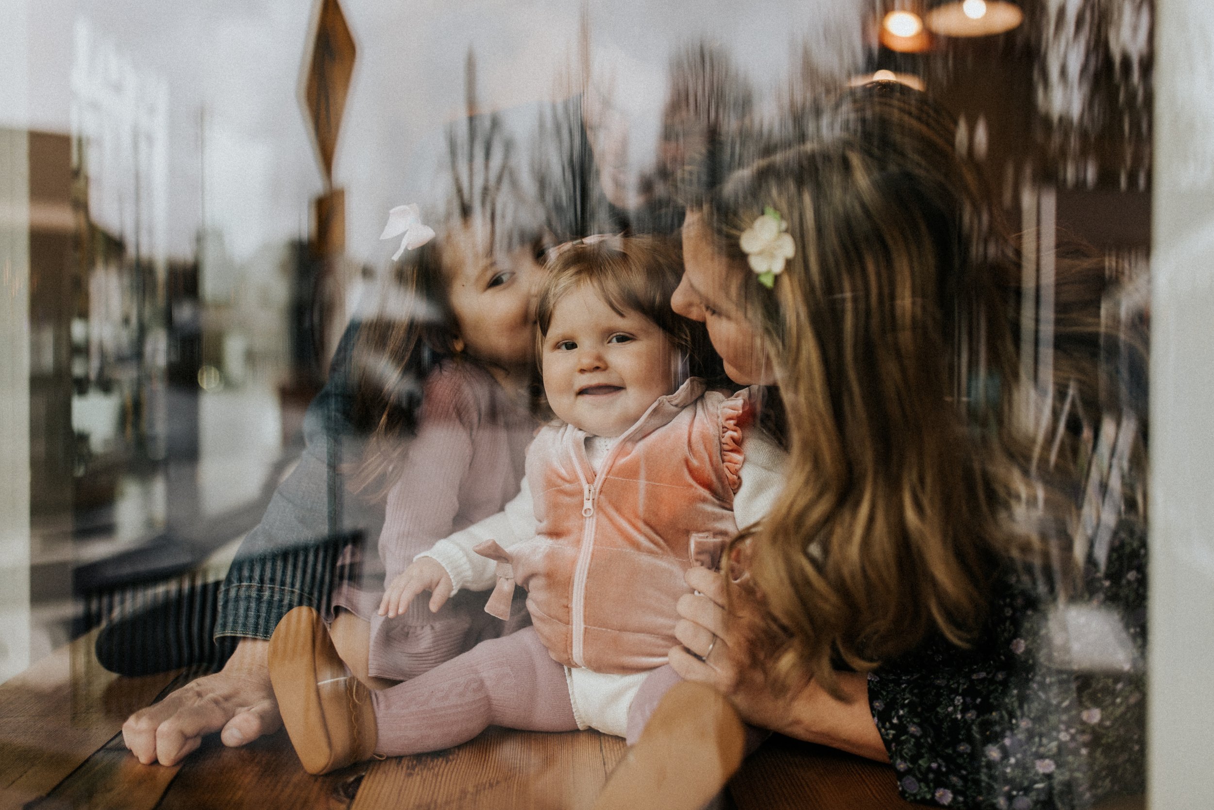 A mother and daughter are sitting in front of a window with a baby in their arms.