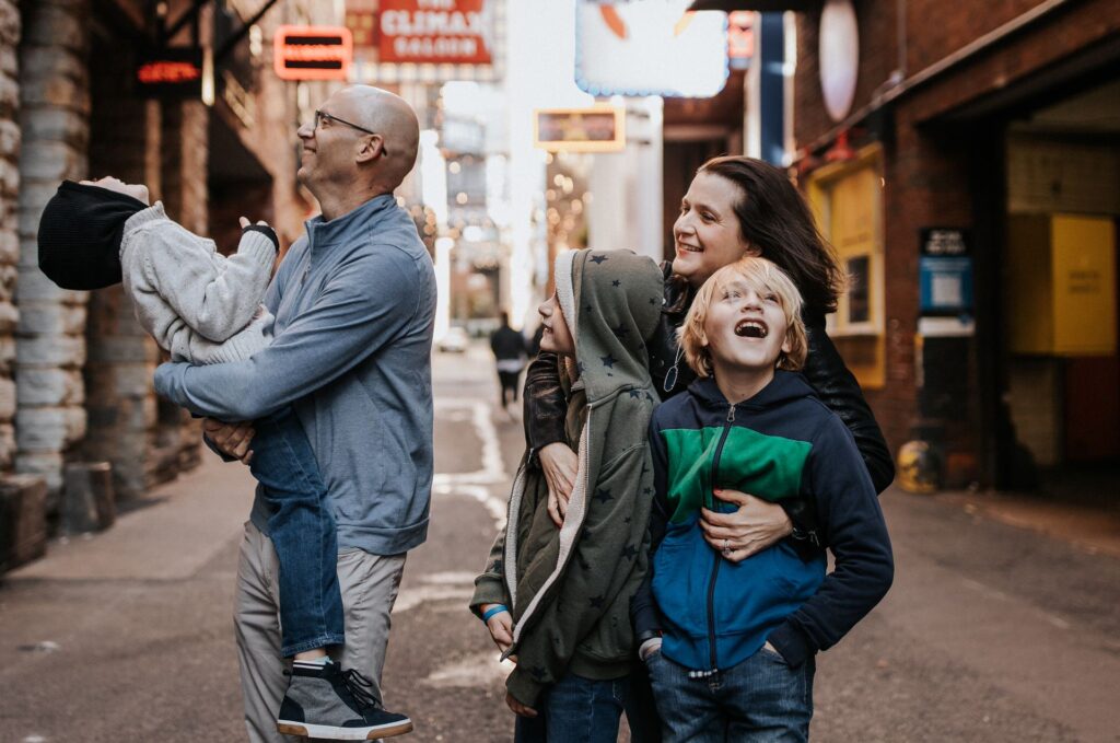 A joyful family of four walking down an urban alley in Portland, with parents holding and interacting with their two young sons.