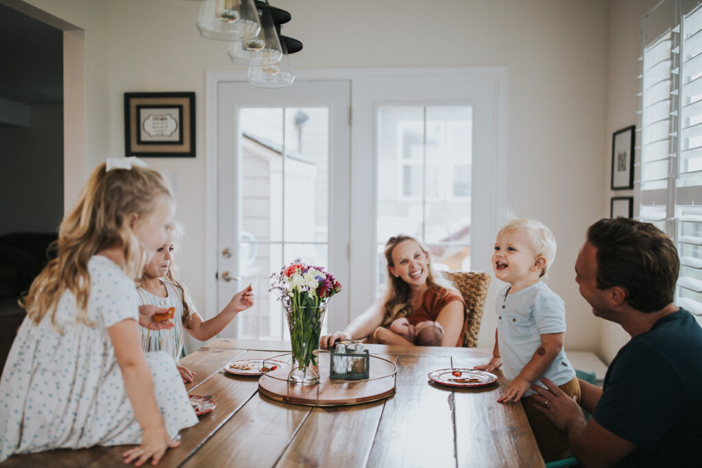 A family of five, including two young children and a baby, enjoys a meal around a wooden dining table adorned with a flower centerpiece in a bright, airy room with large windows—a perfect setting for an impromptu family photoshoot.