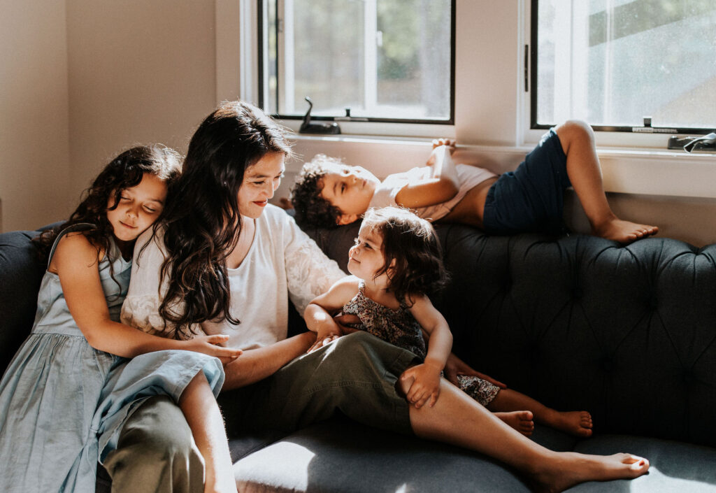 A woman sits on a couch with three children during a family photoshoot; one leans on her shoulder, another snuggles beside her, and the third rests against the window. Sunlight streams through two windows behind them, capturing a perfect moment of warmth and togetherness.