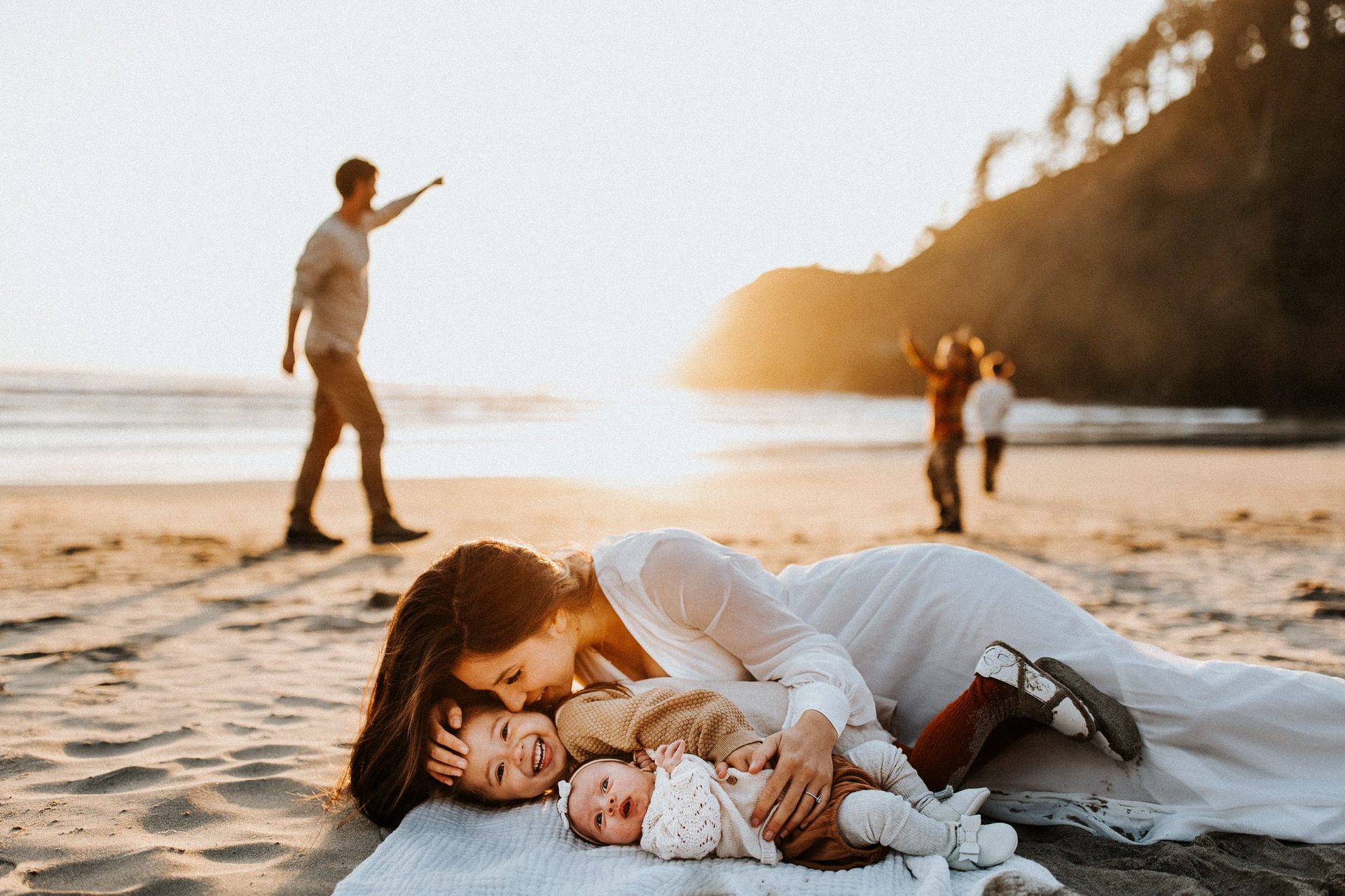 A woman lies on a blanket at the beach with a young child and a baby, while two people walk and point in the background at sunset, capturing the perfect moment for a family photoshoot.