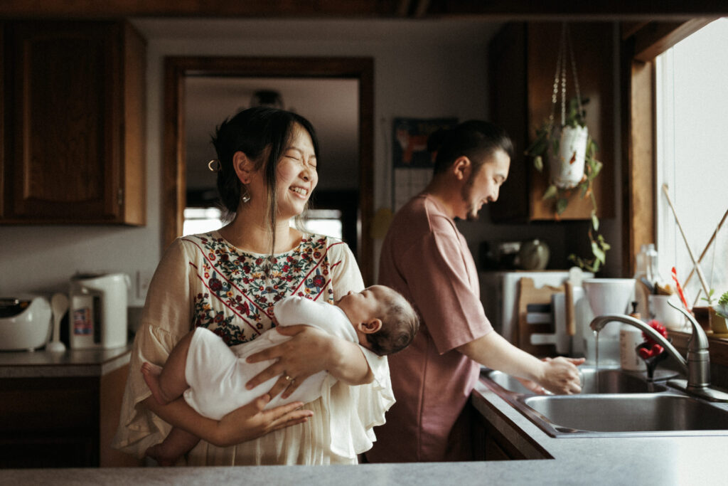 A woman holds a baby and smiles while a man washes dishes in the background. The setting is a kitchen with wooden cabinets and a hanging plant, capturing the intimate moment perfectly that any Portland newborn photographer would cherish.