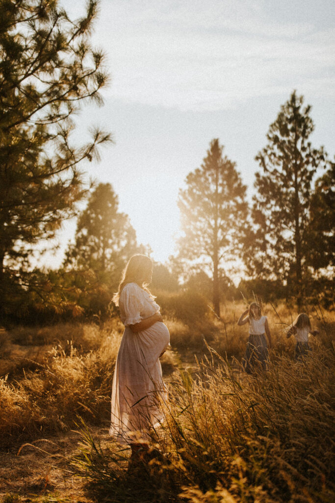 A pregnant woman stands in a sunlit field, surrounded by tall grass and pine trees, captured beautifully by a Portland maternity photographer. Meanwhile, two children play joyously in the background.